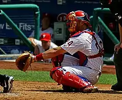 A man in a white baseball uniform with red stripes and wearing a red catcher's mark, red shinguards and cleats, and a light-brown catcher's mitt