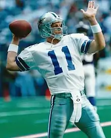 White American male holding a football in throwing position, wearing a silver helmet with a star on the sides and a white jersey and silver pants