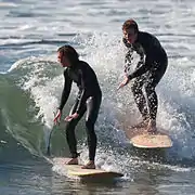 Two surfers wearing one-piece wetsuits, riding a wave.