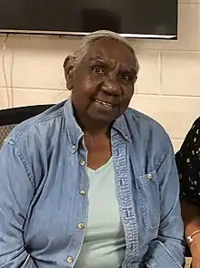 An Aboriginal Australian woman in a denim jacket sits in front of a white wall