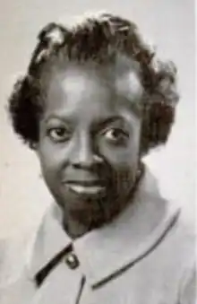 A Black woman with dark skin and hair, wearing a buttoned-up light-colored jacket with a pointed collar