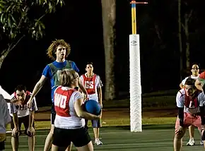 Woman holding a netball while other players stand watching her. Woman and shooting team is wearing white with red bibs. Opposing team is wearing blue with purple bibs.