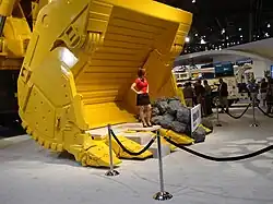 a female model, in a red top and black skirt, standing (with her hands on her hips) in a (bright yellow) bucket of an hydraulic shovel in the exhibit hall of MINExpo 2008. The model is standing behind a velvet rope discouraging trade show attendees from standing in the bucket. Another exhibit is visible in the background as is a group of approximately 12 attendees all but one of which appear to me men.