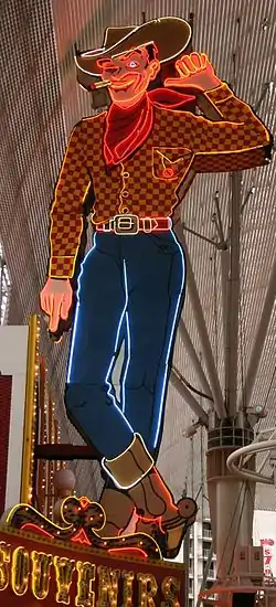 Photograph of a large painted sign in the form of a cowboy. The cowboy is winking his eye. His left hand is lifted, and he's pointing that thumb towards the building to his right. A lighted cigarette dangles from the corner of his mouth. He's wearing a cowboy hat, boots, and a scarf. Glowing neon tubes highlight the outlines.