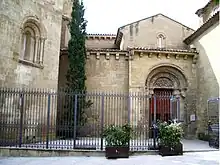 Low yellow-grey stone building behind iron railings, with a decorative door.