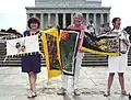 Monica Willard, Justine Merritt, and Michele Peppers meet on the event's tenth anniversary. Merritt is holding two of the Ribbon panels she embroidered.