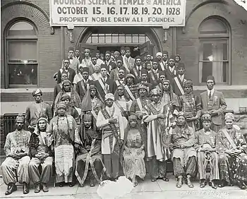 A group of men and women facing forward, dressed in Arabic clothing, assembled outside a building