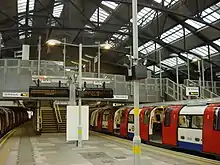 A train, with its red sliding doors open, waits at the right side of a platform. Steps lead up from the platform to a metal bridge crossing the tracks and a partly glazed roof arches on steel trusses high above.