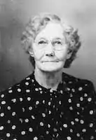 headshot of a white older woman with grey curly hair and glasses wearing a dark shirt with white spots