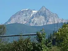 A rocky mountain with little snow at its summit overlooking hilly vegetated terrain in the foreground.