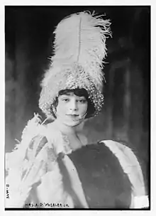 A white woman with dark hair, wearing a high plumed white hat embellished with pearls, and holding a fur muff