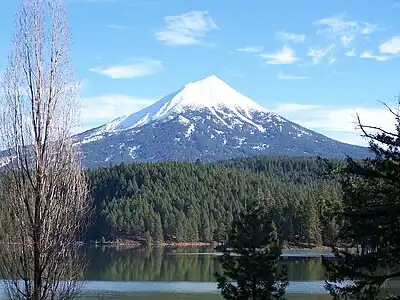 A conical, snow-capped mountain rises above a forest and a lake.