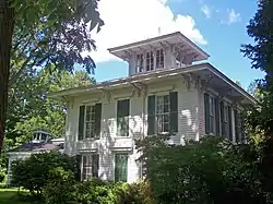 A square white house with green window shutters, wide overhanging eaves and a small square tower in the middle of the roof seen from a corner angle