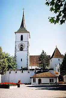 A white church tower with a pointed spire and a dark roof surrounded by a white crenelated wall.