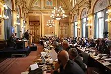 a large baroque yellow and gold room with a stage on the left and long tables filled with men and women in suits on the right.