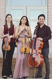 Three musicians holding violin, viola and cello stand together with smiling faces.