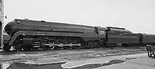 A black-and-white image of a streamlined steam locomotive with its two tenders along with a building in front it on the left