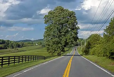 A two-lane road stretching out ahead of the camera to a distant curve to the right. On the left is a rolling of fields, woods and two small houses with a brown fence running alongside the road