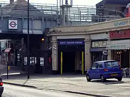 A beige building with a rectangular, dark blue sign reading "NORTH HARROW STATION" in white letters all under a light blue sky with white clouds