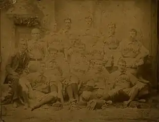 A sepia photograph of 14 men arranged in two rows, standing and sitting. One is wearing a suit while the rest are wearing light baseball uniforms with "Nashville" on the chest and dark socks.