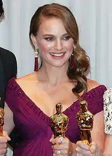 A young woman with brown hair dangling to her left in a V-neck purple dress holding one of the golden statuettes seen above