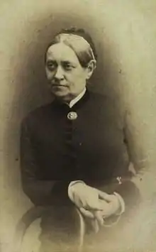 Black and white photo of a woman leaning with her right forearm on the back of a wooden chair