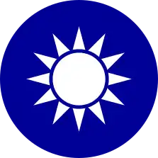 A blue circular emblem on which sits a white sun composed of a circle surrounded by 12 rays.