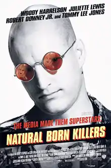 In black-and-white, a bald man sporting a leather jacket wears red sunglasses that contain the reflection of a blonde woman on both lenses.