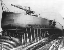 The hull of a large ships in a shipyard, surrounded by scaffolding and cranes