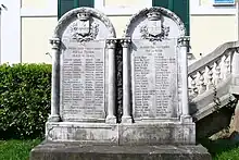 A color photo of two stone memorials, each with approximately 6 feet tall. The WW2 memorial stands on the left, with 46 names inscribed, and the WWI memorial adjoins it on the right, with 42 names inscribed.