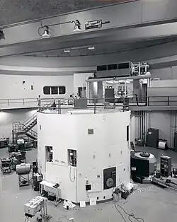 The black and white photograph is of a large room that contains a lot of electronic equipment. The lower half of the image contains a cylindrical white container that is a nuclear reactor. There is a walkway at the top of the reactor, which leads back to a control room where two men are sitting.