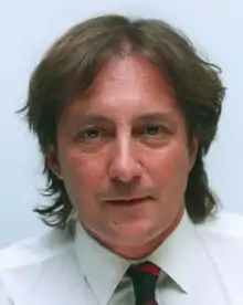 Middle-aged man with shoulder-length brown hair, wearing a white dress shirt, and a necktie with blue and red diagonal stripes