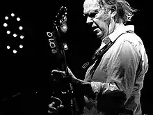 Black and white image of an older man with sideburns wearing a white dress shirt with his sleeves rolled up, holding a guitar.