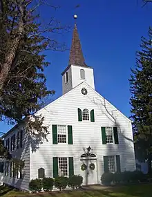  A white building with green shutters and pointed roof. At the front is a pointy tower with a round ball and weathervane on top.