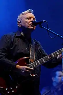 British singer Bernard Sumner singing into a microphone over a blue background, wearing a dark long-sleeved shirt.