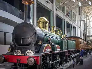 A black, re, and gold-trimmed locomotive with coal tender and two passenger cars sits on a rail siding at the museum