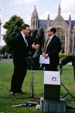 Two men in suits, stood on a grassy field in front of a Gothic style building. There is a tree on the left side, and microphone and recording equipment in the foreground on the floor.