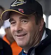 A head and shoulders photograph of Nigel Mansell wearing a black baseball cap with a red number 5 and the subject smiling at the camera