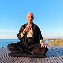 bald, middle-aged White male dressed in black monk-like attire, sitting on black cushion on wood platform next to shoreline, holding right hand up to chest, squinting with a slight smile at camera