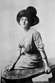 A young woman, seated on a bench, wearing a large dark hat, a light-colored blouse with a scooped neckline, and a light-colored skirt