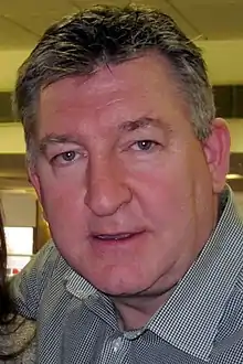 "Colour photograph of a man aged around 40, with grey-brown, wavy hair, wearing a checked grey shirt and looking closely into the camera"