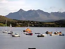 A body of water in the foreground contains several sailing vessels, including yachts, small fishing boats and an orange lifeboat. In the middle distance a variety of modern cottages are set amongst coniferous trees on a long grassy slope. Large and precipitous black mountains dominate the background.