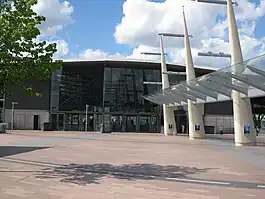 A grey building with grey lampposts in front and a green tree in the foreground all under a light blue sky with billowy, white clouds