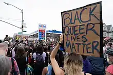 A cardboard protest sign is held with the words: "Black Queer Lives Matter" in the foreground, with hundreds gathering in the background at a Mobil gas station