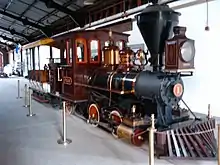 A red steam locomotive with a 0-4-2T wheel arrangement (no leading wheels, four driving wheels, and two trailing wheels) and no tender, coupled to a small train car