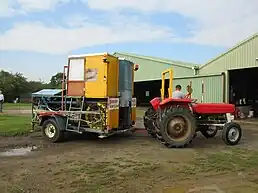 The image shows a tractor on the right with a large, reverse "L"-shaped object attached. The object is the winch, and is constructed from steel frame work and has a wooden cockpit on the top part. It is a mix of blue, yellow and orange colours.