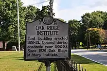 A small sign near a fence containing text. The text reads: OAK RIDGE INSTITUTE First building erected 1851-1852 Opening during academic year 1852–1853. Since 1929, Oak Ridge Military Institute