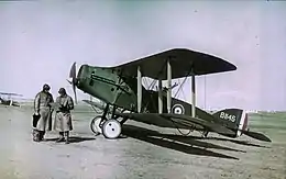 Two men in long leather coats and leather helmets stand in front of a biplane in RAF colours.