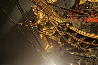 An old-gold coloured figurehead, still attached to the brown wooden ship, of a crowned, bearded man with a double-handed spear. He is covered on his forward arm and just below his waist by flowing cloth. With one foot back towards the ship and one foot forward on gold scroll-work at the bow, he shown starting a charge forward, aiming his spear ahead.