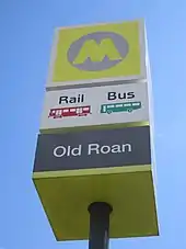 A station sign with a large yellow 'M' logo and the name 'Old Roan' underneath. Icons mark rail and bus services.